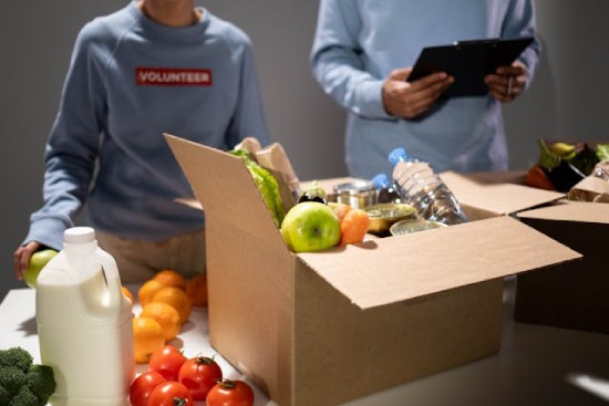 Box of food items in front of two food bank volunteers Volunteers fill boxes with produce, water, and other staples.