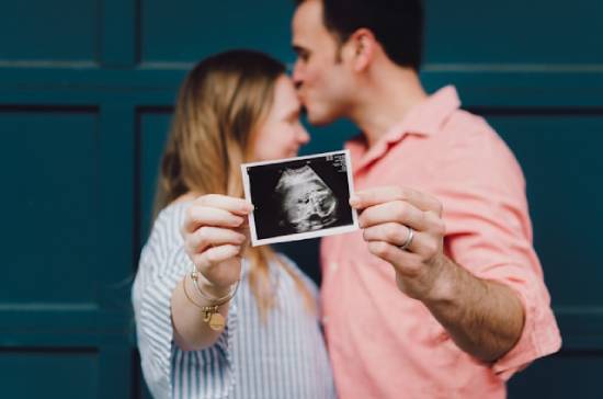 Husband kissing wife while holding an ultrasound picture of their baby A couple in the background happily holds up a sonogram in the foreground.