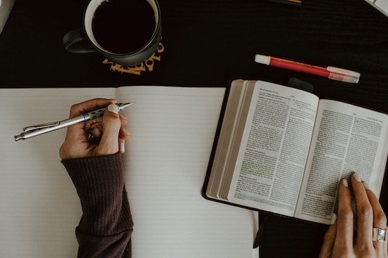 Studying Scripture A woman's left hand sits on top of an open notebook with a pen, ready to write; her right hand rests on an open Bible