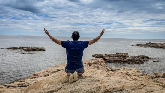 A man kneeling on a rocky shore, hands raised to the sky in repentance.