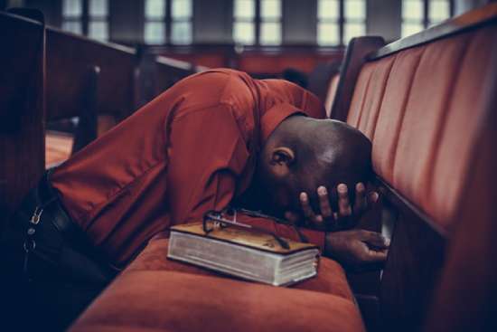 A Man Praying Kneeling and Leaning on Chair A man kneels and prays in a church pew with a Bible beside him, illustrating when we realize we need spiritual healing.