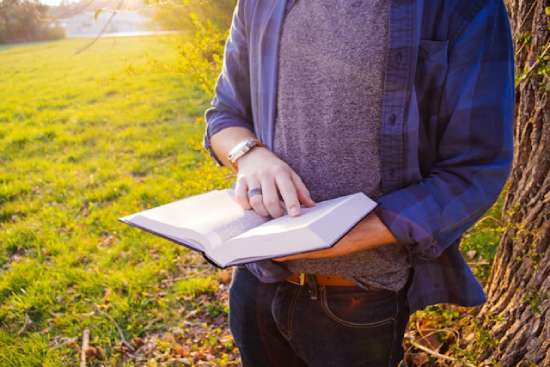 Man Resting Reading Bible A person stands outside, holding an open Bible. Their finger is pointing to a verse that they seem to be considering.