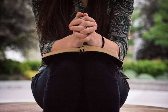 Simple Prayer Position Woman sitting while praying with folded hands on an open Bible as we learn about various physical postures during prayer