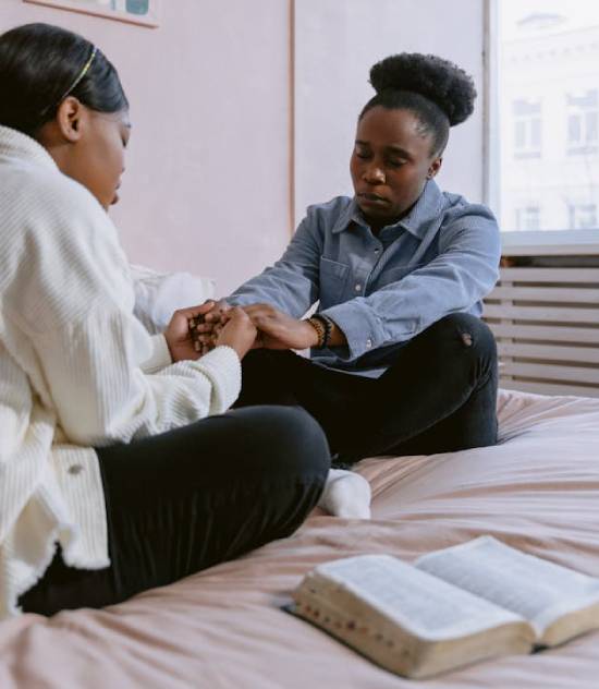 Two Women Praying while Sitting on a Bed Two women sitting on a bed pray together, giving us an example of how prayer can comfort us when we feel discouraged.