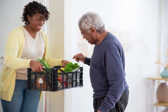 Woman holding a crate of vegetables in front of an elderly man who is choosing what he likes A young woman happily offers a basket of produce to an elderly man.