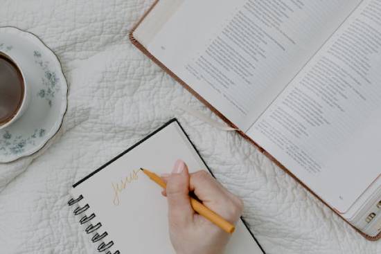 Woman's Hand Writing Jesus' name in a Notebook Beside an Open Bible and Cup of Tea A woman sits on a bed with her morning coffee, an open Bible, and a notebook. She writes "Jesus" at the top of a page, taking note of what she's learning about Him as she studies the Word.
